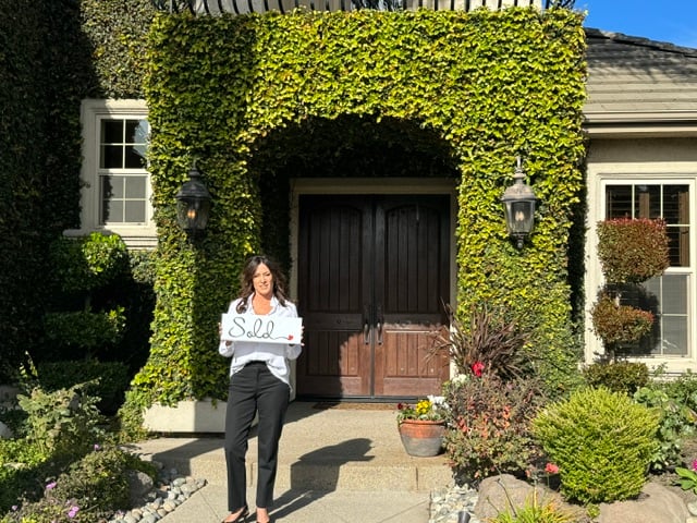 Audrey Schmierer smiling and holding a Sold sign on the front walk of an ivy-covered home with wooden double doors.