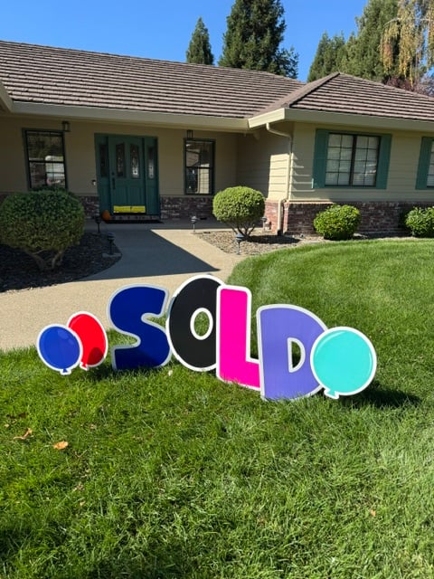 A colorful SOLD yard sign with balloons on the front lawn of a single-story home with a green front door