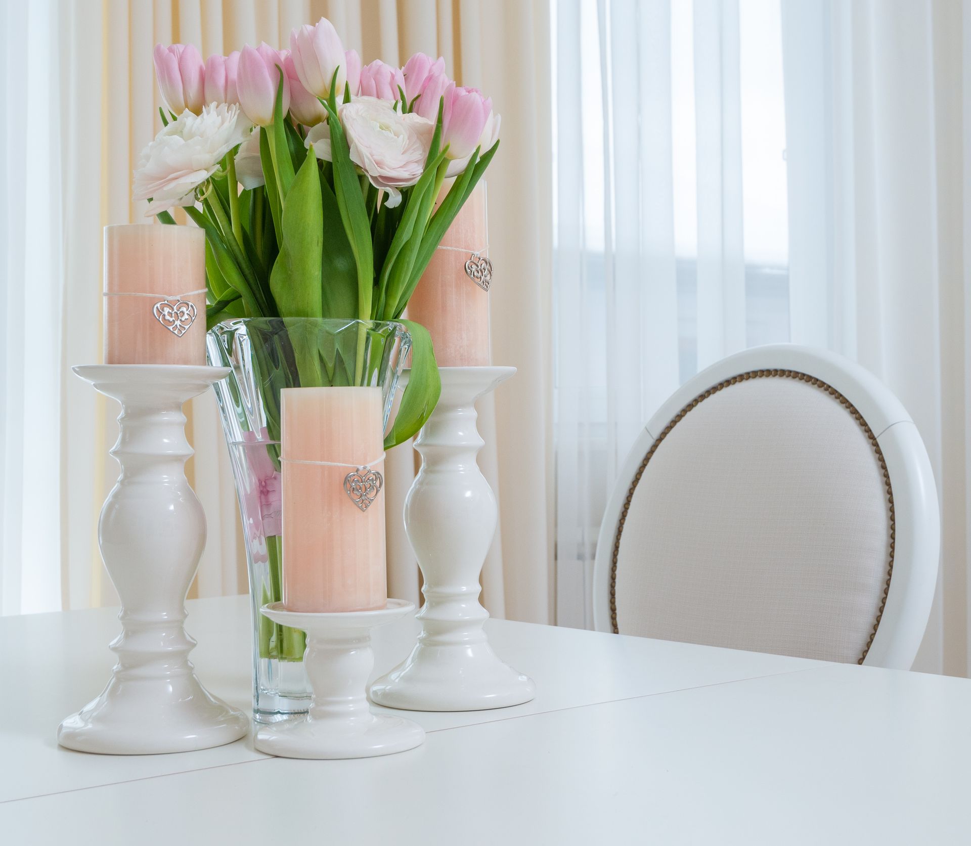 Pink tulips and white flowers in a glass vase with three peach candles on a white dining table, beside a white upholstered chair.